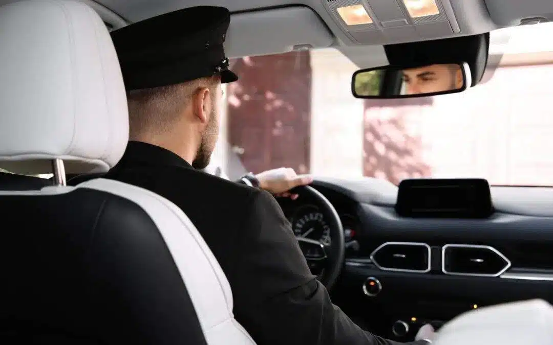 A chauffeur in a black uniform and hat sits in the driver’s seat, his face visible in the rearview mirror, ready to provide a VIP Fast Track Transfer. The modern car interior features white seat covers and sleek dashboard controls.
